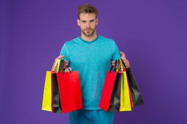 Alışveriş torbaları menekşe arka plan üzerinde olan adam. Maço renkli kağıt torbalar ile. Moda shopper rahat mavi tshirt. Tatil hazırlanması ve kutlama. Alışveriş veya satış ve cyber Pazartesi.