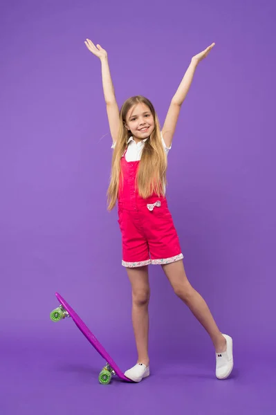 幼年期およびアクティブなゲーム 小さな女の子は 紫色の背景にスケート ボードと笑顔します 気ままな一日 ロングボードを浮かべて子供スケーター ピンクのジャンプ スーツ子供のスケート ボード スポーツ活動とエネルギー — ストック写真