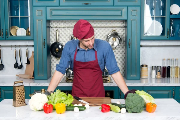 Cook in chef hat and apron at kitchen table. Cook man with vegetables and tools ready for cooking dishes.