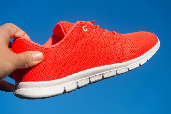 Sneaker on blue sky. Sneaker in hand on sunny outdoor. Sport shoe of orange fabric material on white sole. Fashion style and trend. Sport and active lifestyle. Run the life