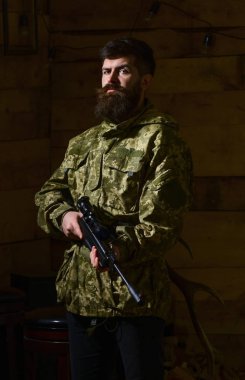Sakallı kamuflaj elbiseler, ahşap iç arka plan giyer. Avcı, acımasız hipster silahla avlamaya hazır. Avcı kavramı. Maço gamekeepers evinde sıkı yüzünde