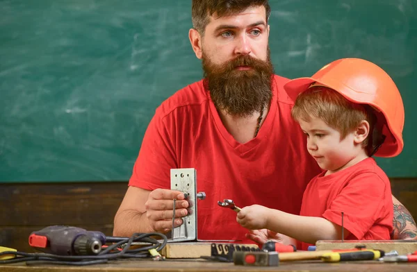 Vater mit Bart lehrt kleinem Sohn Werkzeug im Klassenzimmer, Kreidetafel im Hintergrund. Männerarbeitskonzept. Junge, Kind im Schutzhelm bastelt von Hand, repariert, bastelt mit Papa — Stockfoto