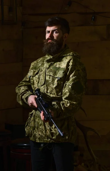 Man with beard wears camouflage clothing, wooden interior background. Hunter, brutal hipster with gun ready for hunting. Huntsman concept. Macho on strict face at gamekeepers house