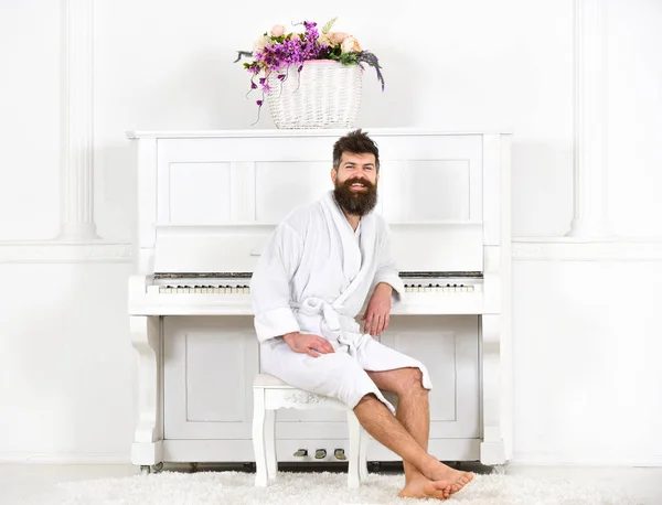 El hombre con barba en albornoz disfruta de la mañana mientras está sentado cerca del piano. El hombre alegre se sienta delante del instrumento musical de piano en el interior blanco sobre el fondo. Concepto de músico talentoso —  Fotos de Stock