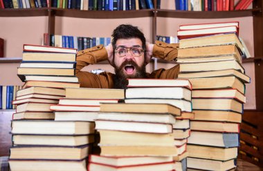Bilimsel bir keşif kavramı. Adam kazık kitap kütüphane, kitap rafı arka plan üzerinde arasında şok yüzünde. Öğretmen ya da öğrenci sakallı gözlük takmaz, defocused kitaplar ile masada oturur