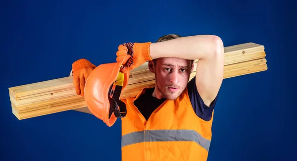 Man in helmet and protective gloves wiping sweat from forehead, blue background. Tired labourer concept. Carpenter, woodworker, labourer, builder on tired face carries wooden beam on shoulder