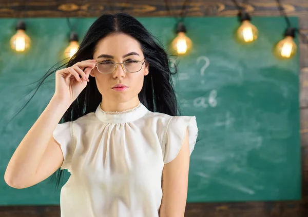 教室で白いブラウスに長い髪の女が立っています。穏やかな顔の女性の厳しい先生は、黒板の前に立ちます。メガネと手を振って髪先生は魅力的に見えます。厳しい先生のコンセプト — ストック写真