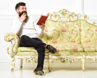 Sakal ve bıyık oturur barok tarzı kanepe, tutar kitap, beyaz duvar arka plan üzerinde olan adam. Adam zevk ile eski kitap okuma. Mizahi edebiyat kavramı. Maço yüz kitap okuma gülüyor üzerinde