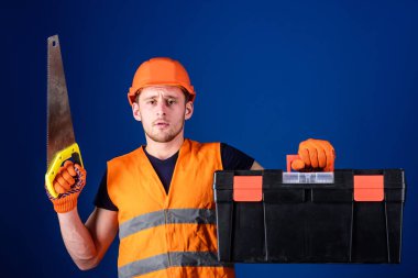 Marangoz kavramı. İşçi, repairer, tamirci üzerinde ciddi bir yüz taşıyan araç, onarım, kopya alanı için hazır. Kask, kask içinde adam araç kutusu taşır ve el testeresi, mavi arka plan tutar
