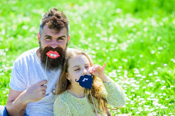 Father posing with lips and child posing with beard photo booth attribute. Gender roles concept. Dad and daughter sits on grassplot, grass on background. Family spend leisure outdoors — Stock Photo, Image