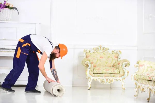 Relocating concept. Man with beard, worker in overalls and helmet rolling carpet, white background. Courier delivers furniture in case of move out, relocation. Loader wrappes carpet into roll