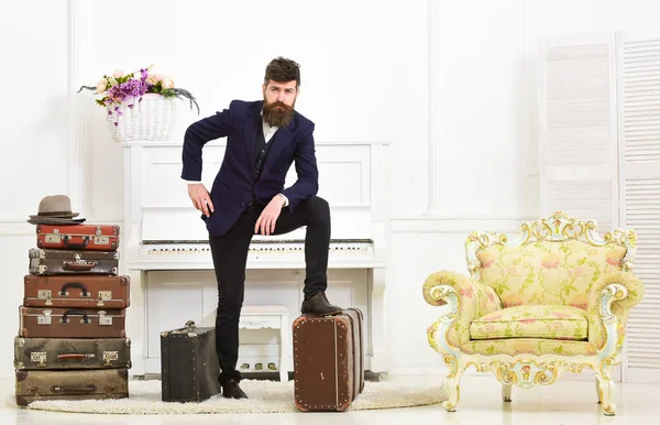 Macho elegant on thoughtful face standing near pile of vintage suitcase. Man, traveller with beard and mustache with luggage, luxury white interior background. Luggage and travelling concept