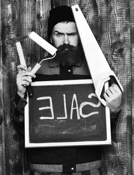 Homem barbudo segurando várias ferramentas de construção e placa, rosto sério — Fotografia de Stock