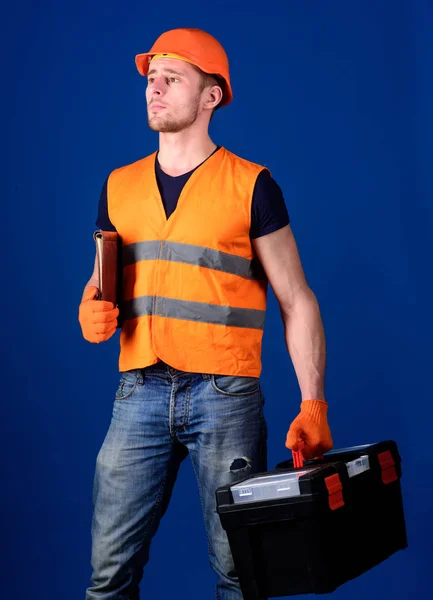 Mann mit Helm, Mütze hält Werkzeugkiste und Ordner mit Dokumenten, blauer Hintergrund. Arbeiter, Reparateur, Bauarbeiter mit verträumtem Gesicht tragen Koffer mit Werkzeugen. Reparateur bereit zur Arbeit. Reparaturdienstkonzept — Stockfoto