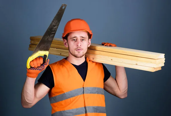 Man, handyman in helmet, hard hat holds handsaw and wooden beams, grey background. Woodcraft concept. Carpenter, woodworker, labourer, builder on thoughtful face carries wooden beams on shoulder — Stock Photo, Image