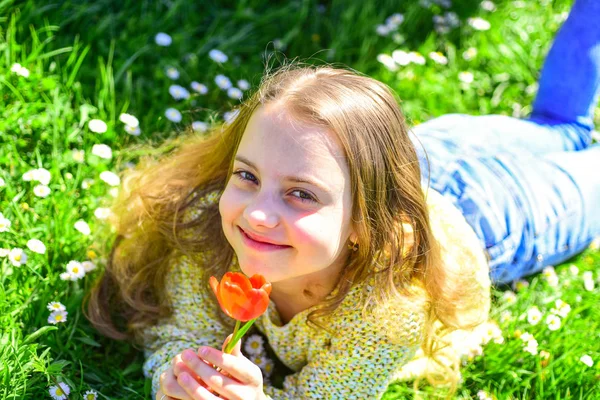 Dívka na usmívající se tvář drží červený Tulipán květ, vychutnat aroma. Mládež a bezstarostné koncepce. Dítě si jarní slunečný den vleže na louce s květinami. Dívka ležící na trávě, grassplot na pozadí — Stock fotografie