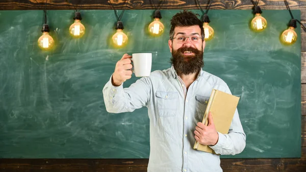 Fika koncept. Mannen med skägg på glada ansikte i klassrummet. Lärare i glasögon håller boken och mugg kaffe eller te. Forskare håller boken och mugg kaffe, svarta tavlan på bakgrunden kopia utrymme — Stockfoto