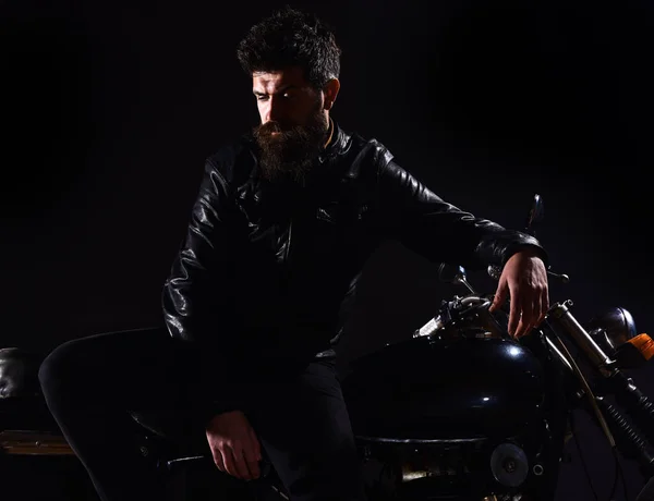 Macho, brutal motociclista con chaqueta de cuero se para cerca de la motocicleta por la noche. Brutalidad y concepto masculino. Hombre con barba, motorista en chaqueta de cuero inclinado en moto en la oscuridad, fondo negro — Foto de Stock