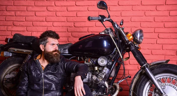 Мужчина с бородой, байкер в кожаной куртке возле мотоцикла в гараже, кирпичный настенный фон. Концепция образа жизни байкеров. Хипстер, жестокий байкер на задумчивом лице в кожаной куртке сидит, опирается на мотоцикл — стоковое фото