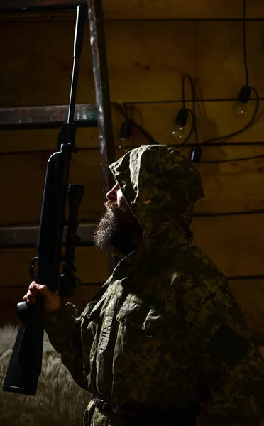 Huntsman concept. Hunter, brutal hipster with gun prepare rifle for hunting. Macho on strict face at gamekeepers house. Man with beard wears camouflage hooded clothing, wooden interior background — Stock Photo, Image