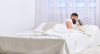 Arka plan üzerinde yatak uyanık, beyaz perde döşeme tişörtlü adam. Yatak odası konsepti. Maço sakal ve bıyık nap sonra rahatlatıcı. Adam sabah uyanma uykulu güler yüz