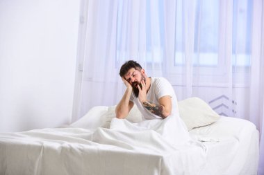 Uykulu yorgun yüz esneme üzerinde adam. Tişörtlü adam yatak, beyaz perde arka plan üzerinde oturur. NAP ve siesta kavramı. Maço sakal ve bıyık esneme, rahatlatıcı, nap, sahip rest