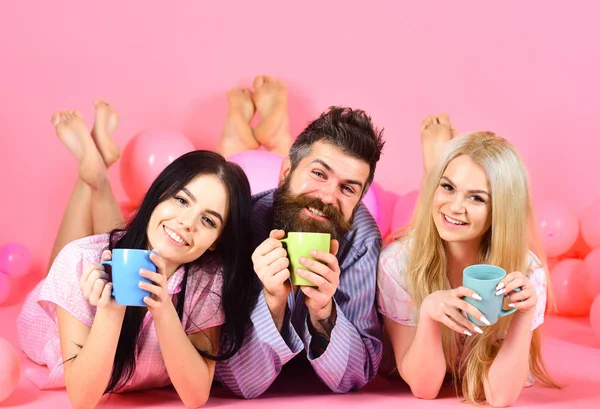 Lovers drinking coffee in bed. Man and women in domestic clothes, pajamas. Threesome relax in morning with coffee. Lovers in bed concept. Man and women, friends on smiling faces lay, pink background — Stock Photo, Image