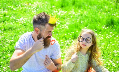Baba ve kızı, grassplot, arka planı yeşil çimenlerin üzerinde oturur. Aile açık havada eğlence harcamak. Çocuk ve babası crown ve gözlük ile poz booth öznitelikleri fotoğraf. En iyi arkadaşlar kavramı