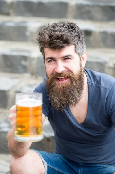 Le hipster barbu tient une tasse de bière, boit de la bière en plein air. Homme avec barbe et moustache sur le visage heureux, fond pierreux, déconcentré. Concept de bière artisanale. Hipster à longue barbe semble détendu — Photo