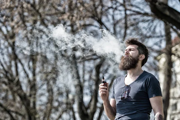 Hombre barbudo fumando cigarrillos electrónicos al aire libre. Un hombre elegante paseando por la ciudad exhalando vapor. El cigarrillo electrónico como alternativa al tabaco —  Fotos de Stock