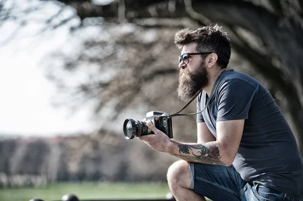 Bearded man works with vintage camera. Man with beard and mustache on concentrated face, branches on background, defocused. Man with long beard busy with shooting photos. Photographer concept — Stock Photo, Image
