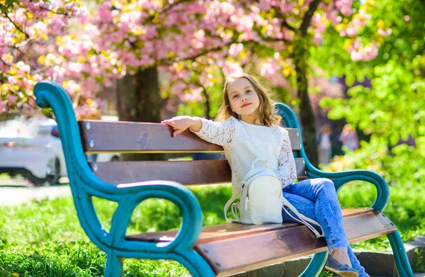 Koncepcja akcesoria. Relaks podczas spaceru w parku, w pobliżu wiśniowy kwiat dziewczyna. Dziewczyna na uśmiechniętą twarz siedzi na ławce, drzewa sakura na rozmytym tłem. Słodkie dziecko z plecaka cieszyć się słoneczny wiosenny dzień — Zdjęcie stockowe