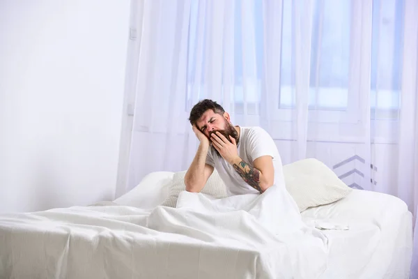 Um tipo com o rosto cansado e sonolento a bocejar. Macho com barba e bigode bocejando, relaxando, dormindo, descansando. Conceito de sesta e sesta. Homem de camisa senta-se na cama, cortinas brancas no fundo — Fotografia de Stock