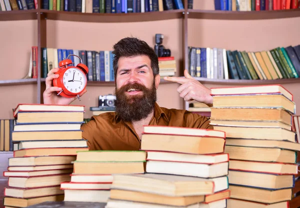 Mann, Wissenschaftler, der mit Wecker aus Bücherstapeln lugt. Lehrer oder Schüler mit Bart studieren in der Bibliothek. Mann mit glücklichem Gesicht zeigt auf Uhr, Bücherregale im Hintergrund. Zeitflusskonzept — Stockfoto