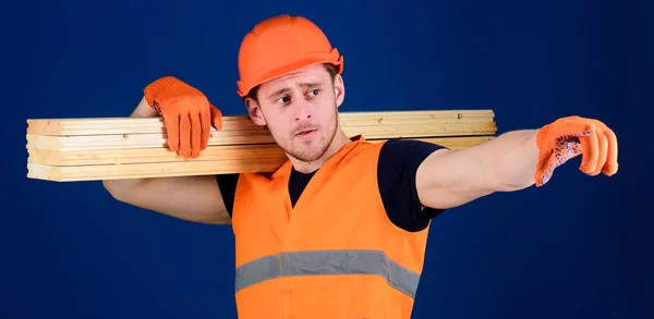 Ο άνθρωπος στο κράνος, κράνος και προστατευτικά γάντια που δείχνει την κατεύθυνση, μπλε φόντο. Ξύλινα υλικά έννοια. Ξυλουργός, ξυλουργός, ισχυρό οικοδόμος στο στοχαστικό πρόσωπο ασκεί ξύλινα δοκάρια στον ώμο — Φωτογραφία Αρχείου