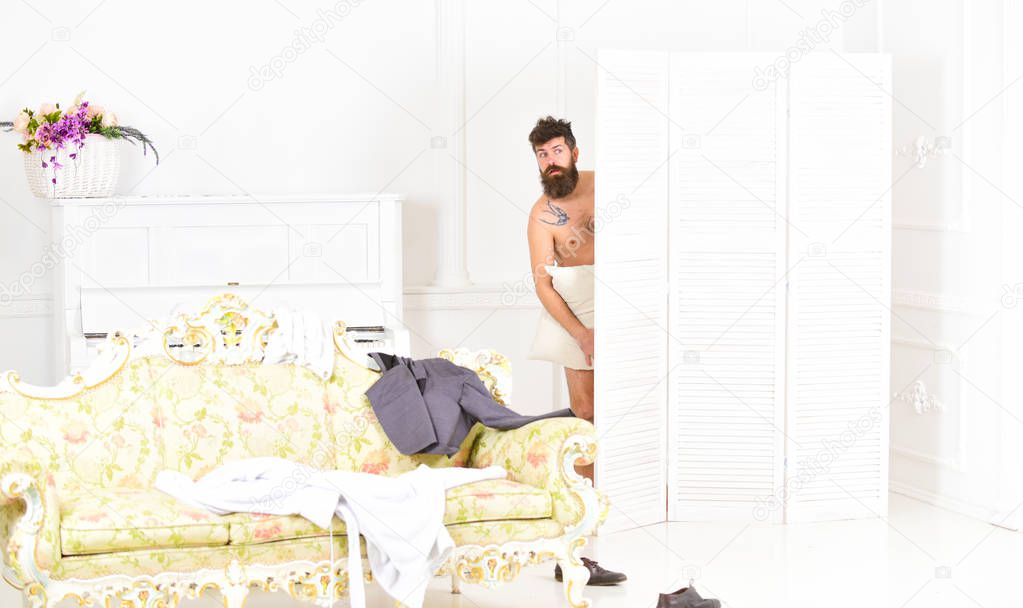 Man, lover in white interior caught naked. Lovers and cheating concept. Hipster naked on shocked face unexpectedly detected in bedroom. Man with beard and mustache hiding behind folding screen