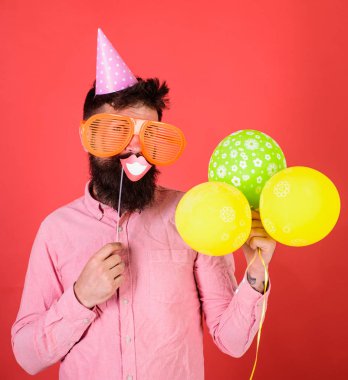 Hippi dev güneş gözlüğü kutluyor. Neşeli yüzündeki sakallı gülümseyen ağız sopa, kırmızı bir arka plan üzerinde tutar. Fotoğraf booth eğlence kavramı. Hava balonları ile parti şapkalı adamı kutluyor