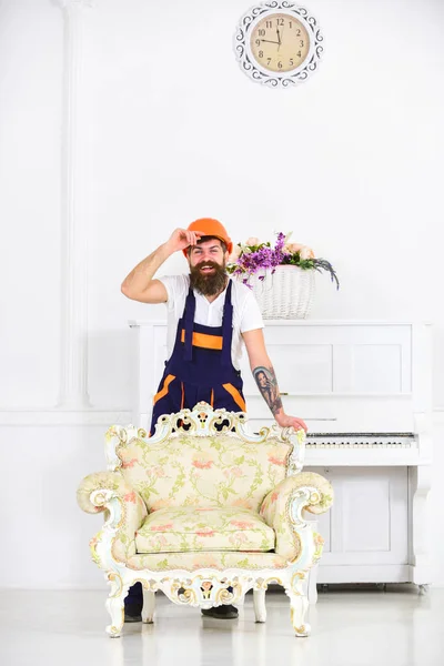 Happy mover de pie detrás de un sillón vintage con estampado florido. Tipo barbudo en casco naranja frente al piano aislado sobre fondo blanco — Foto de Stock