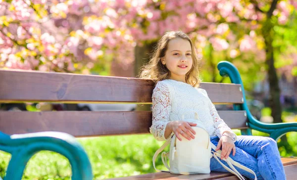 背景には、デフォーカスの桜の木のベンチに座っている笑顔の女の子。アクセサリーのコンセプトです。桜の近くの公園で散歩しながらリラックスした少女。バックパックでかわいい子は、日当たりの良い春の日をお楽しみください。 — ストック写真