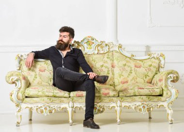 Hipster kibirli yüzünde yalnız oturuyor. Sakallı ve bıyıklı lüks oturma odasında boş zaman harcıyor. Zengin ve yalnız kavramı. Lüks daire sahibi beyaz duvar arka koltukta oturur