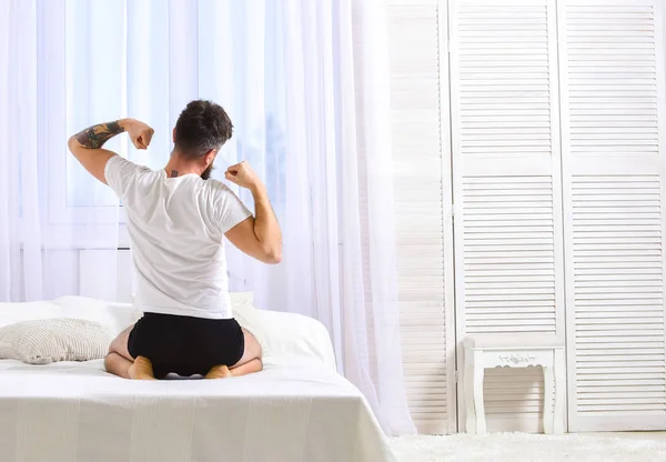 Guy stretching arms, full of energy in morning, rear view. Perfect morning concept. Man in shirt sits on bed, white curtains on background. Macho in underpants stretching, relaxing after nap, rest