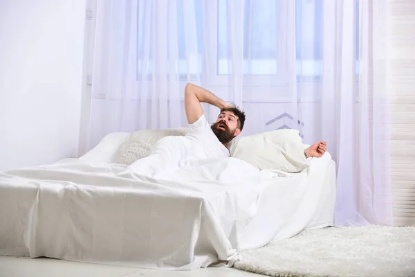 Hombre de camisa acostado en la cama despierto, cortina blanca en el fondo. Un tipo con la cara sorprendida despertando por la mañana. Macho con barba y bigote se quedó dormido despertando. Despertar y dormir concepto —  Fotos de Stock