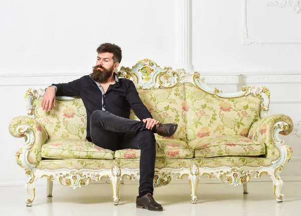 Hipster mit arrogantem Gesicht sitzt allein. Mann mit Bart und Schnurrbart verbringt Freizeit im Luxus-Wohnzimmer. reiches und einsames Konzept. Besitzer der Luxuswohnung sitzt auf Sofa, weißer Wandhintergrund — Stockfoto