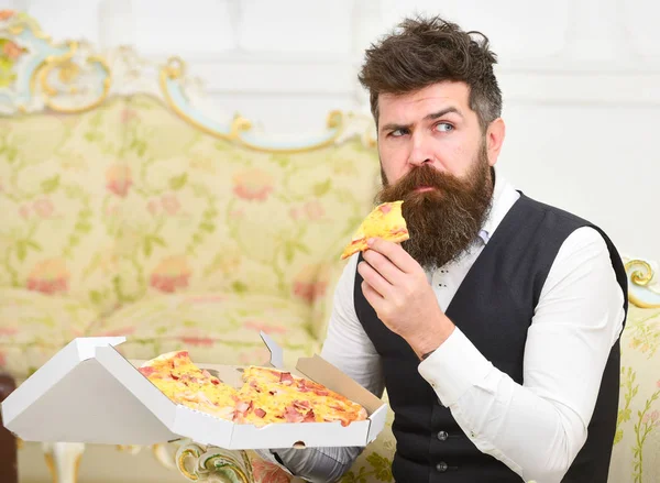 Man with beard and mustache holds delivered box with tasty fresh hot pizza. Macho in classic clothes hungry, on strict face, holds slice of pizza, eats, interior background. Pizza delivery concept