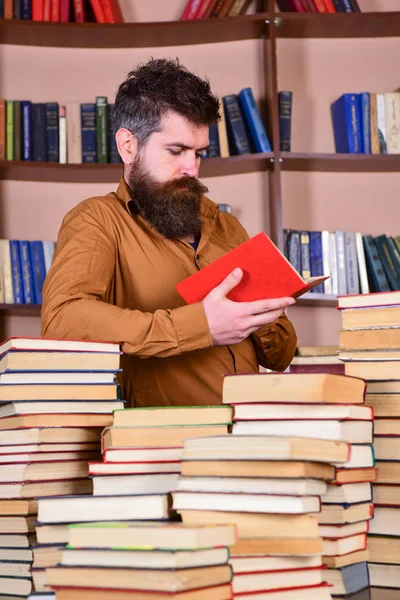 Образование и наука. Учитель или ученик с бородой учится в библиотеке между грудами книг. Человек на занятом задумчивом лице читает книгу, книжные полки на заднем плане — стоковое фото