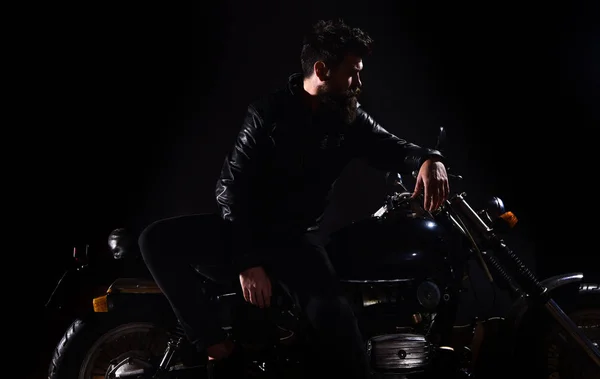 Man with beard, biker in leather jacket lean on motor bike in darkness, black background. Biker culture concept. Macho, brutal biker in leather jacket stand near motorcycle at night time, copy space