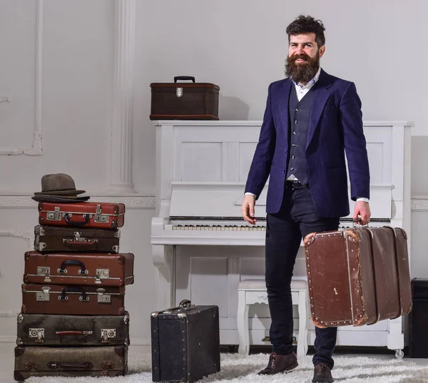 Macho elegant auf lächelndem Gesicht steht neben einem Stapel alter Koffer, hält Koffer. Mann, Reisender mit Bart und Schnurrbart mit Gepäck, luxuriöser weißer Innenraum. Gepäckausgabekonzept — Stockfoto