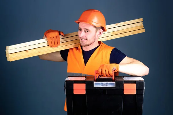 Concetto professionale di falegname. Falegname, operaio, costruttore, falegname sul volto sorridente porta travi in legno sulla spalla. Uomo in casco, cappello rigido porta attrezzi e travi in legno, fondo grigio — Foto Stock