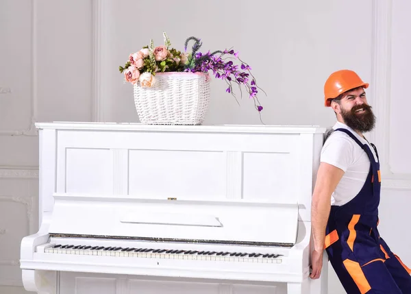 El mensajero entrega muebles, se muda, se traslada. Hombre con barba trabajador en casco y overoles empuja, poner esfuerzos para mover el piano, fondo blanco. Concepto de cargas pesadas. Cargador se mueve instrumento de piano — Foto de Stock