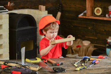 Miğferli şirin çocuk inşaatçı ya da tamirci olarak oynuyor, tamir ediyor ya da el işi yapıyor. El işi konsepti. Meşgul suratlı çocuk atölyede evde oynuyor. Çocuk tahtaya çivi çaktı.